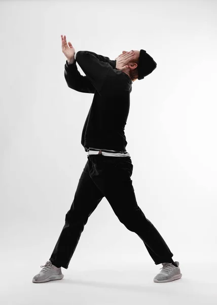 Jovem dançarina estilo livre vestida com jeans pretos, camisola, chapéu e tênis cinza está dançando fechando seu rosto com a mão no estúdio no fundo branco — Fotografia de Stock