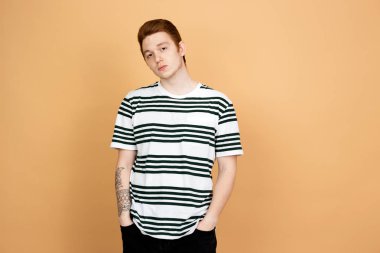 Çizgili gömlek ve siyah kot pantolon kırmızı saçlı adam stüdyoda bej arka planda ceplerinde elleri ile duruyor