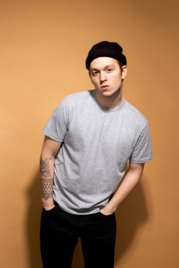 Young stylish guy dressed in a gray shirt, black jeans and black hat with tattoo on his hand is posing on the beige background in the studio