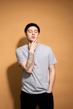 Young stylish guy dressed in a gray shirt, black jeans and black hat with tattoo on his hand is posing on the beige background in the studio