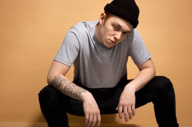 Ukala şık adam gri gömlek giymiş, siyah kot pantolon ve elinde dövme ile siyah şapka stüdyoda bej arka plan üzerinde çömelme olduğunu