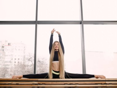 Spor kıyafetleri giymiş uzun sarı saçlı güzel atletik kız jimnastik salonunda ahşap pencere üzerinde böler oturuyor