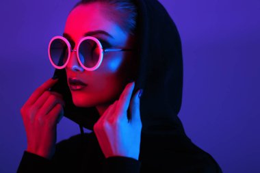 Siyah kazaklı bir kukuleta ile ve güneş gözlüğü kırmızı ve mavi neon ışık stüdyo yuvarlak genç kız moda portresi