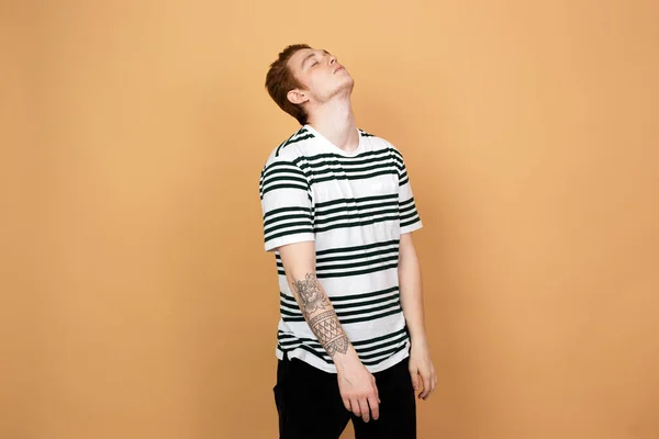 Red-haired stylish guy in a striped shirt and black jeans with tattoo on his hand stands on the beige background in the studio