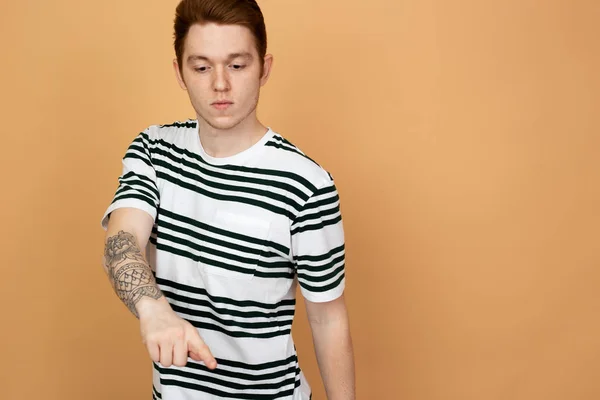 Red-haired stylish guy in a striped shirt with tattoo on his hand is posing on the beige background in the studio