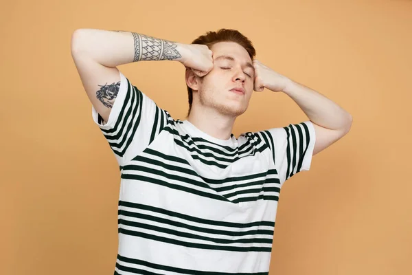 Red-haired stylish guy in a striped shirt with tattoo on his hand is posing holding his hands on his head on the beige background in the studio