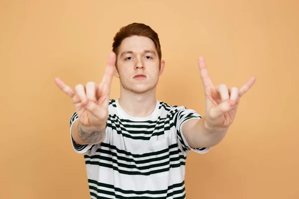 Homme élégant aux cheveux rouges dans une chemise rayée avec tatouage sur la main est montre panneau rack avec ses mains sur le fond beige dans le studio — Photo