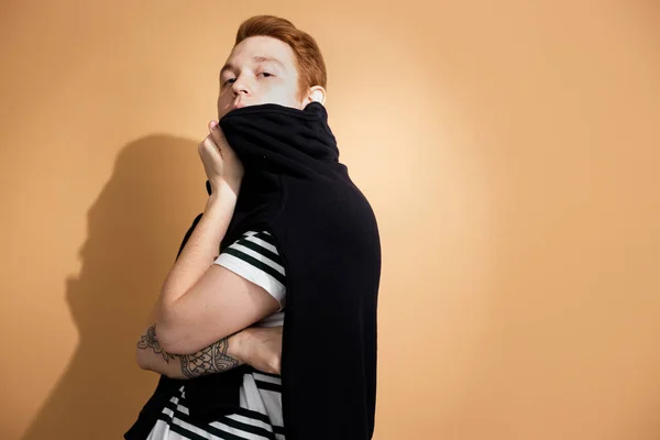 Stijlvolle roodharige jongen in een gestreepte shirt met tatoeage op zijn hand is poseren met zwarte trui op zijn schouders op de beige achtergrond in de studio — Stockfoto