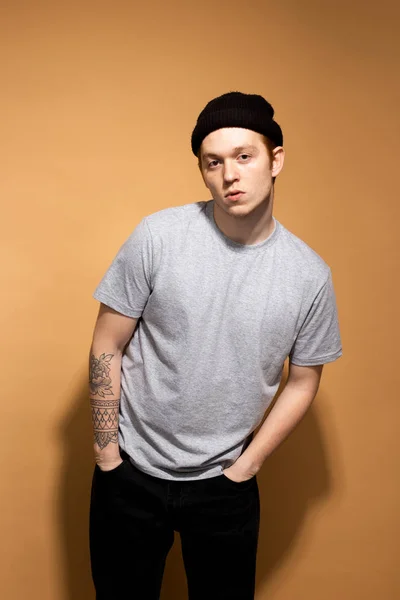 Young stylish guy dressed in a gray shirt, black jeans and black hat with tattoo on his hand is posing on the beige background in the studio — 스톡 사진