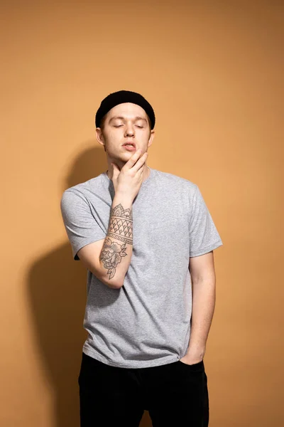 Young stylish guy dressed in a gray shirt, black jeans and black hat with tattoo on his hand is posing on the beige background in the studio — Stockfoto