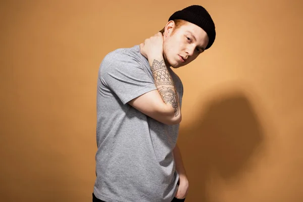 Young stylish guy dressed in a gray shirt and black hat with tattoo on his hand is posing on the beige background in the studio — Stockfoto