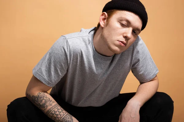 Nafoukaný chlapík oblečený do šedé košile, černé džíny a černý klobouk s tetováním na ruce se Krká na béžové pozadí studia — Stock fotografie