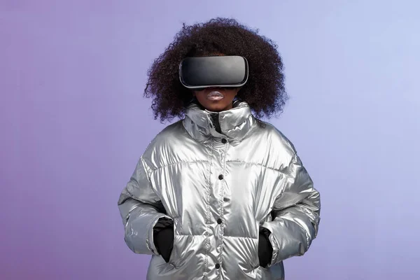 Moderna chica de pelo castaño rizado vestida con una chaqueta de color plateado utiliza las poses de gafas de realidad virtual en el estudio sobre fondo de neón — Foto de Stock