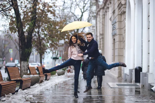 Casal feliz, cara e sua namorada vestida com roupas casuais estão pulando sob o guarda-chuva na rua na chuva . — Fotografia de Stock