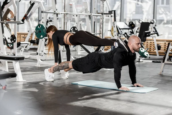 Sportliches Paar bei schwierigen Sportübungen, wo das Mädchen in der Planke auf einem Mann steht, der in der Planke in der modernen Turnhalle steht — Stockfoto