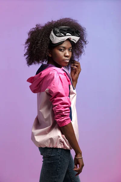 Mod curly brown-haired girl dressed in the pink sports jacket wearing on her head the virtual reality glasses poses in the studio on neon background — Stock Photo, Image