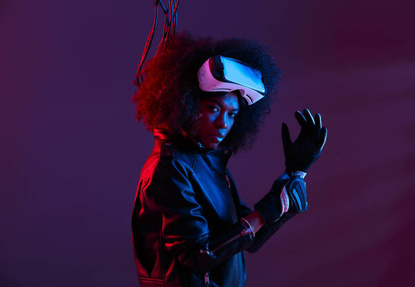 Curly dark haired girl dressed in a black leather jacket and gloves is wearing  the virtual reality glasses on her head in the dark studio with neon light
