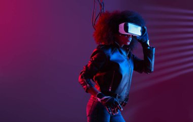 Siyah deri ceket ve eldiven giymiş kıvırcık saçlı bir kız karanlık stüdyoda kafasında neon ışıklı sanal gerçeklik gözlüğü kullanıyor.