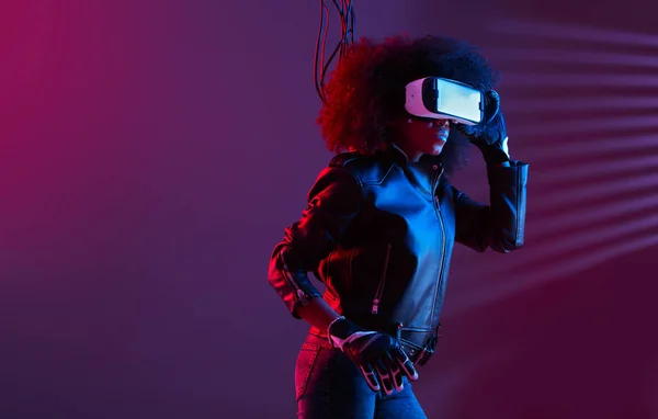 Mod chica de pelo oscuro rizado vestido con chaqueta de cuero negro y guantes utiliza las gafas de realidad virtual en su cabeza en el estudio oscuro con luz de neón — Foto de Stock