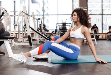 Spor kıyafetler giymiş güzel ince kıvırcık koyu saçlı kız ekipman bir sürü modern spor salonunda fitness için mat üzerinde oturuyor
