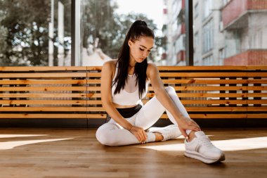 Beyaz spor kıyafetler giymiş ince koyu saçlı kız spor salonunda pencerenin yanında yere oturur