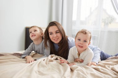 Mutlu bir aile. Açık mavi pijama giymiş genç anne büyük pencere ile yatak odasında bej battaniye ile yatakta onu iki küçük oğlu ile yatıyor