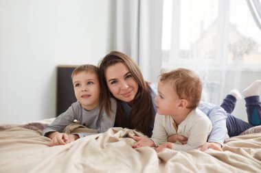 Mutlu bir aile. Açık mavi pijama giymiş genç anne büyük pencere ile yatak odasında bej battaniye ile yatakta onu iki küçük oğlu ile yatıyor