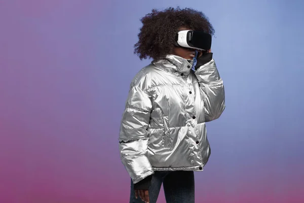 Chica de pelo castaño rizado de moda vestida con una chaqueta de color plateado utiliza las gafas de realidad virtual en el estudio sobre fondo de neón — Foto de Stock