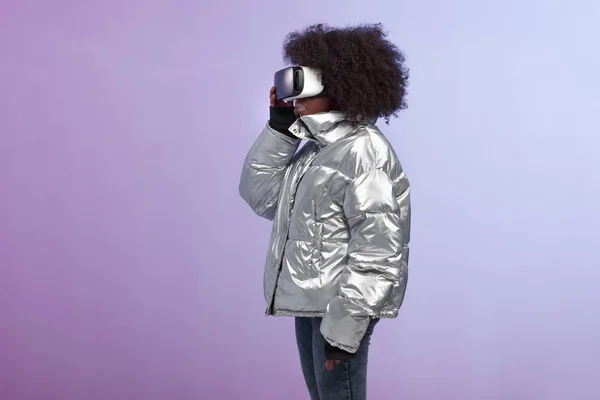 Chica de pelo castaño rizado de moda vestida con una chaqueta de color plateado utiliza las gafas de realidad virtual en el estudio sobre fondo de neón — Foto de Stock