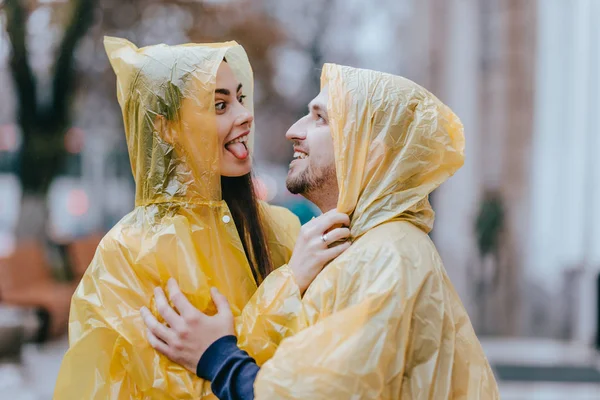 Feliz casal amoroso, cara e sua namorada vestida com capas de chuva amarelas estão abraçando na rua na chuva — Fotografia de Stock
