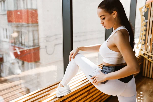 Giovane ragazza dai capelli scuri sottile vestita con abiti sportivi bianchi fa stretching vicino alla finestra in palestra — Foto Stock