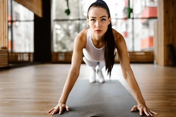 Güzel ince koyu saçlı kız beyaz spor giysiler giyinmiş spor salonunda tahta yapıyor yapıyor