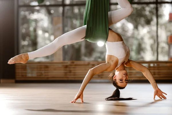 Belle fille mince aux cheveux foncés habillée en haut de sport blanc et collants fait des exercices d'étirement sur hamac vert — Photo