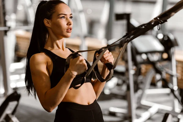 Siyah spor üst ve şort giymiş atletik koyu saçlı kız spor salonunda fitness istasyonu üzerinde çalışıyor — Stok fotoğraf