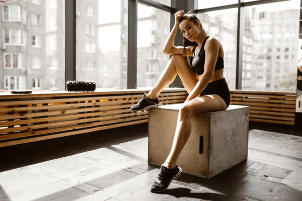Charmant laster meisje gekleed in zwarte sporttop en shorts zit op een houten kist in het zonlicht aan de voorkant van het raam in de sportschool — Stockfoto
