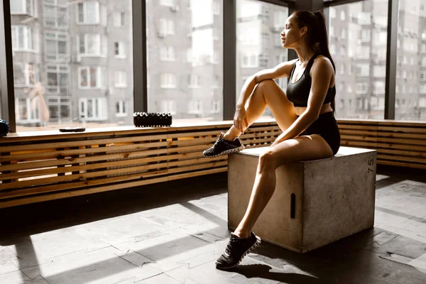 Чарівна дівчина, одягнена в чорний спортивний топ і шорти, сидить на дерев'яній коробці на сонячному світлі перед вікном у спортзалі — стокове фото
