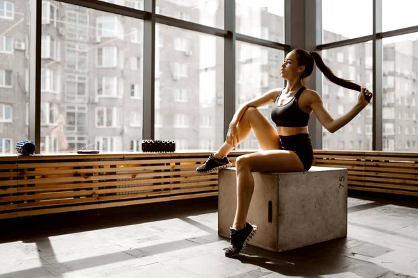 Mooi slank meisje gekleed in zwarte sporttop en shorts zit op een houten kist in het zonlicht aan de voorkant van het raam in de sportschool — Stockfoto
