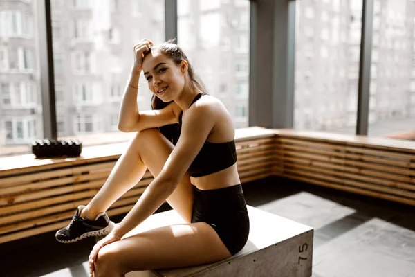 Hermosa chica delgada vestida con top deportivo negro y pantalones cortos está sentada en una caja de madera a la luz del sol en frente de la ventana en el gimnasio — Foto de Stock