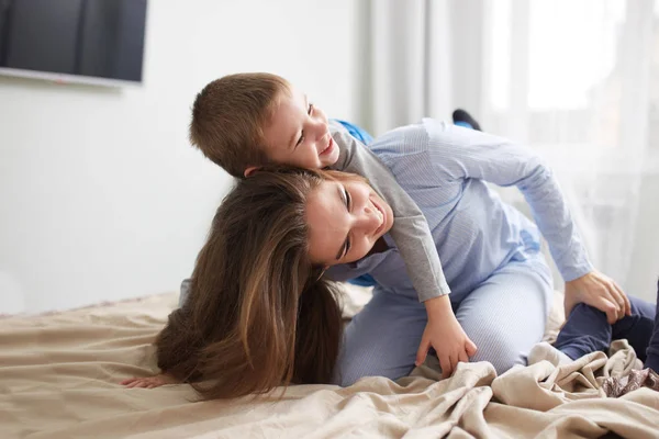 Młoda piękna matka i jej synka, ubrana w piżamy zabawy na łóżku w sypialni światło przytulny — Zdjęcie stockowe