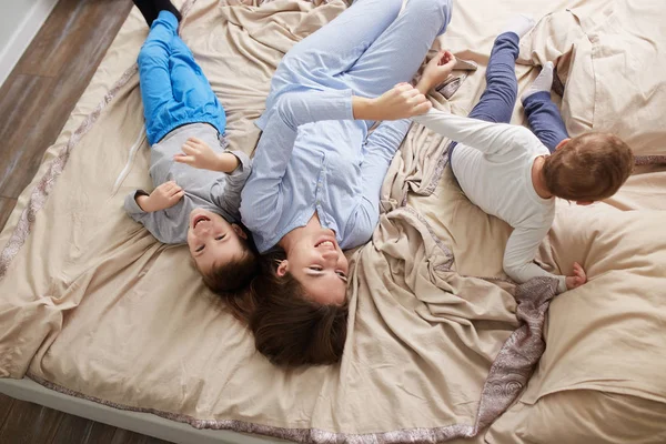 Щаслива красива мати, одягнена в світло-блакитну піжаму, лежить зі своїми двома маленькими синами на ліжку з бежевою ковдрою в спальні — стокове фото