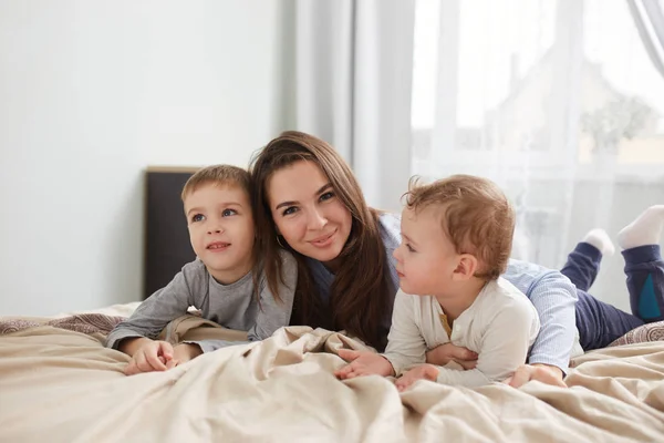 Szczęśliwa rodzina. Młoda matka ubrana w jasnoniebieską piżamę, z jej dwoma małymi synami na łóżku z beżowym kocem w sypialni z dużym oknem — Zdjęcie stockowe