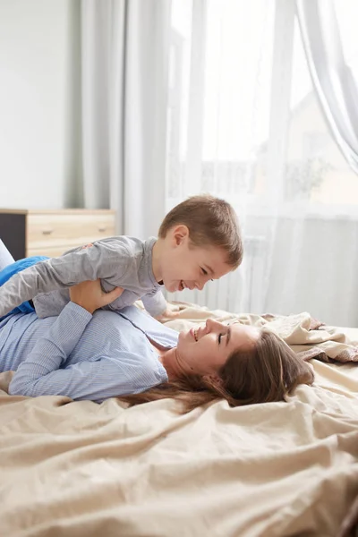 Açık mavi pijama giymiş genç bir anne bej bir battaniyeyle yatağa uzanıyor ve küçük oğlunu büyük pencereli yatak odasında tutuyor. — Stok fotoğraf