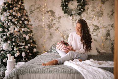 Beyaz kazak ve pantolon giymiş koyu saçlı kız elinde bir Yeni Yıl ağacı ve mumlar ile dekore edilmiş bir odada gri battaniye ve beyaz yastıklar ile yatakta oturan bir Yeni Yıl hediyesi tutar