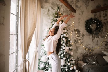 Beyaz kazak ve pantolon giymiş Büyüleyici kız pencerenin önünde Yeni Yıl ağacının yanında duruyor ve Yeni Yıl çelenk ile rahat bir dekore odada uzanır