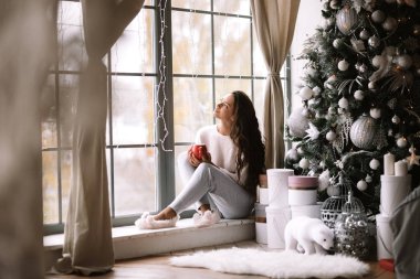 Pantolon, kazak ve sıcak terlik giymiş Büyüleyici koyu saçlı kız yeni yıl ağacı, hediyeler ve mumlar yanında odada panoramik bir pencerenin pencere üzerinde oturan kırmızı bir fincan tutar
