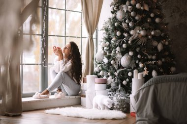 Pantolon, kazak ve sıcak terlik giymiş Büyüleyici koyu saçlı kız yeni yıl ağacı, hediyeler ve mumlar yanında odada panoramik bir pencerenin pencere üzerinde oturan kırmızı bir fincan tutar
