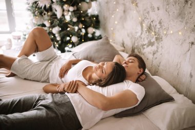 Mutlu adam ve kız beyaz t-shirt giymiş gri yastıklar ile beyaz bir battaniye ile bir yatakta yatıyor ve Yeni Yıl ağacının yanında el ele tutuşuyor