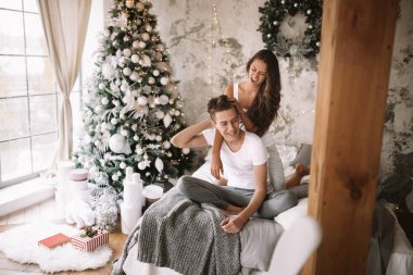 Mutlu adam ve kız beyaz t-shirt giymiş oturma ve yeni yıl ağacı, hediyeler ve mumlar ile rahat bir dekore odada gri bir battaniye ile yatakta sarılma