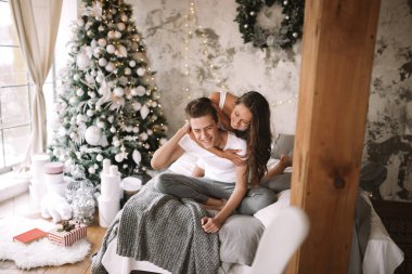 Mutlu adam ve kız beyaz t-shirt giymiş oturma ve yeni yıl ağacı, hediyeler ve mumlar ile rahat bir dekore odada gri bir battaniye ile yatakta sarılma