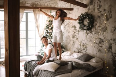 Mutlu kız beyaz t-shirt ve şort giymiş bir Yeni Yıl ağacı, hediyeler ve mumlar ile rahat bir dekore odada oturan adamın yanında yatakta atlıyor